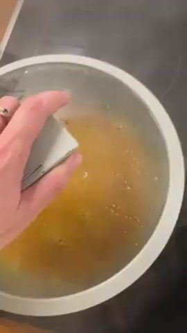 A person lifts a transparent lid from a pot sitting on a black stovetop. The pot contains boiling water and pasta. Steam rises from the pot. The stovetop has a digital timer display.