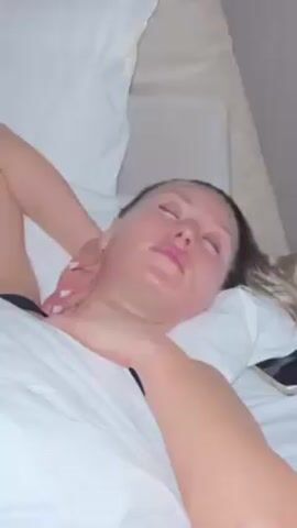 A woman with fair skin lies on her back in a bed covered with white sheets. Her eyes are closed and her face appears slightly red. She has light brown hair and pink lips. One of her arms is bent and resting on her chest while the other is on top of her neck. A hand is seen massaging her neck. A smartphone is visible on the bed beside her. The environment suggests an indoor setting.