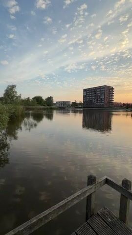 The video shows a serene scene of a lake reflecting buildings and a partly cloudy sky at sunset. A wooden dock is visible in the foreground, with the water surface having slight ripples. On the opposite side of the lake, there is a tall, dark building with multiple windows, flanked by lower residential structures. The sky has a mix of blue and orange hues from the setting sun, casting a warm glow on the scene. The overall composition is calm and picturesque.