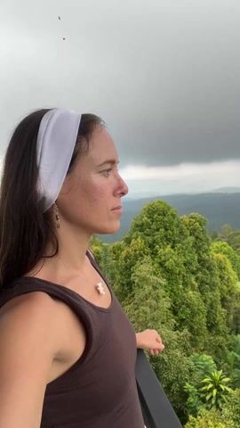A medium shot of a woman in a brown tank top and a white headband, standing on a balcony with a view of a green, hilly landscape under a cloudy sky. She is looking to the side, then turns to face the camera and smiles slightly. She is wearing earrings and a necklace. The balcony has a dark railing. Tropical plants, including banana trees, are visible below the railing.