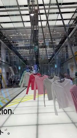 The video shows an indoor display of clothing. A row of pastel-colored long-sleeved shirts are hanging on display racks in what appears to be a retail environment. The shirts are arranged in a line, moving away from the camera and towards the background. The shirts are predominantly in shades of pink, white, light gray, and a pale green. The display floor is illuminated with white light tiles, creating a grid pattern. Behind the clothing display, a mirrored wall reflects the scene, creating a sense of depth and symmetry. In the distance, blurred figures of people can be seen, suggesting the presence of shoppers or staff. The ceiling is composed of white panels with visible lighting fixtures. The overall impression is a modern and clean retail space with an emphasis on the clothing presentation.