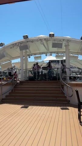 A band plays music on a stage with a white canopy. The band is comprised of three people, one of whom is singing into a microphone. The stage is elevated and accessible by a set of wooden stairs. The area in front of the stage is a wooden deck with chairs. The sky above is clear and blue, suggesting a sunny day. There are lights and speakers mounted on the canopy. The scene appears to be outdoors, possibly on a cruise ship or at a resort.