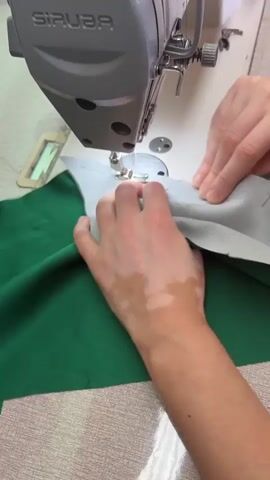 A person's hands are shown sewing two pieces of fabric together with a gray SIRUBA sewing machine. The fabric on the left is dark green and shiny, while the fabric on the right is light gray. The person's right hand holds the fabric steady, and their left hand guides it under the needle. The person's left hand has patches of vitiligo. The sewing machine is on a light-colored surface, possibly a table. The background is slightly blurred.