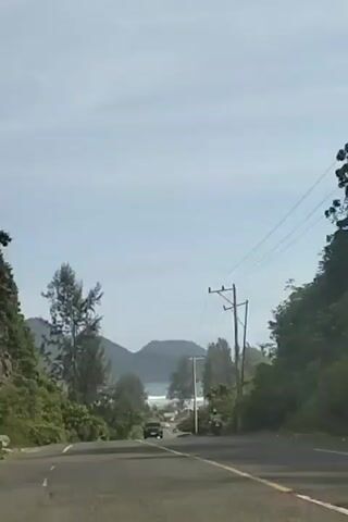 A black car is driving on a road. A motorcycle is also driving on the road. The road is lined with trees and power lines. In the distance, there are mountains and the ocean.