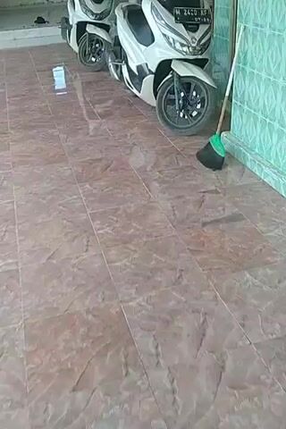 Two white scooters are parked on a tiled floor. The scooter in the foreground is a Honda PCX 160, with a license plate reading M 2420 KF. A broom with a green handle and black bristles leans against the wall next to the scooter. The scooter in the background is partially visible and appears to be identical to the one in the foreground. The floor is covered in reddish-brown tiles with a marble-like pattern. The wall is light green with a textured pattern.