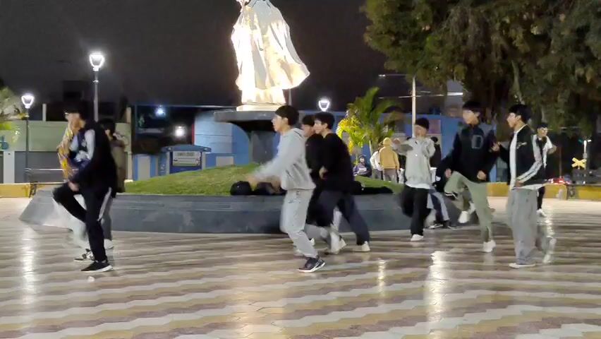 The video shows a group of about 10 young men dancing in a public square at night. They appear to be practicing a choreographed routine, moving in unison and breaking out in jumps and coordinated steps. The square is lit by streetlights and features a statue of a religious figure on a raised platform in the center.  Several people are seen seated on the platform behind the statue, watching the dancers. The square has a patterned tile surface, and is bordered by buildings and trees. The dancers are dressed in casual clothing, including sweatpants, hoodies, and jackets.