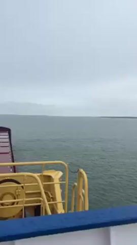 The camera shows a view from a boat on a gray, overcast day.  The water is choppy.  Land is visible in the distance. Parts of the boat's infrastructure, painted yellow and blue, are visible in the foreground.  A dark, rectangular light fixture is bolted to the boat's railing on the right.  As the camera moves, a gray cover also becomes visible.