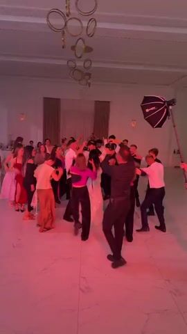 A wedding reception is underway with guests dancing in a large, brightly lit room. A photographer is positioned in the center of the dance floor, holding a professional camera and a large flash unit. The lighting in the room shifts between pink, blue, and green hues. Several guests are captured in motion, some with their arms raised, enjoying the music and celebration. The room appears to be decorated with elegant chandeliers and draped curtains.