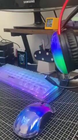 The video shows a desktop computer setup with a Samsung monitor on a wooden shelf, with a lit-up keyboard, mouse, and headset on a gridded mat. There's also a Rubik's cube, a small device with a digital display, and a mushroom-shaped object used as a pen holder. The peripherals have rainbow LED lights changing color. A yellow digital clock shows the time. The shot is static and close-up, focusing on the illuminated desk items.