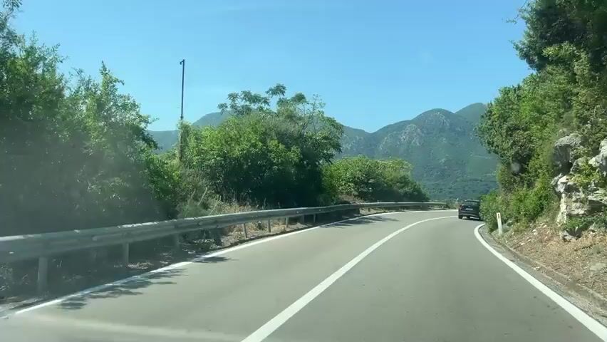 The video shows a first-person perspective from a vehicle driving on a paved road. The road has white lane markings and a metal guardrail on the left side. Lush green trees and vegetation line the road, with mountains visible in the background under a clear blue sky. The sun appears to be bright. A black car is visible in the distance on the right side of the road. The camera view is from inside the vehicle, likely from the dashboard or windshield. The road appears to be winding.