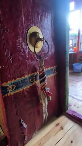 A close-up shot of a dark red, wooden door shows a brass door knocker with a circular metal ring attached. A braided cord with white, black, and colorful stripes hangs from the ring, along with some off-white fabric tassels. The door is decorated with a horizontal band of blue, yellow, and dark green painted designs. The bottom of the door has a small metal latch. To the right of the door, a wooden floor is visible, and a partially open doorway reveals a room with a wooden floor, a blue bucket, and a shelf with orange and white containers. Natural light streams in from the right.