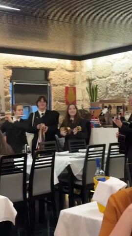 The video shows a group of four young people in dark jackets performing music in a restaurant. Three of them are visible in full frame, while one person is partially visible on the far left. The person second from the left is playing a flute or a similar instrument and also holding a tambourine. The person to their right is playing a ukulele. The person on the far right is playing a guitar. All four are wearing dark blazers with what appear to be ties or ribbons. They are standing behind tables with white tablecloths, and several chairs are visible. The restaurant has stone walls and a tiled ceiling. There are decorations on the wall, including what looks like a painting. A bottle of wine is visible on one of the tables. The lighting is warm and appears to be from overhead fixtures. The video is shaky and appears to have been filmed by someone moving around.