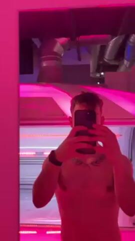 A person stands in front of a mirror, taking a selfie with a smartphone. The room is illuminated with bright pink and white lights, which are reflected in the mirror. The background shows a room with a tanning bed, which the person then enters. The camera moves abruptly as they step inside, showing the interior of the tanning bed with its lights and gridded surface.