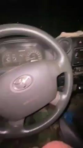 Here is a description of the video:

The video shows the interior of a Toyota Land Cruiser, focusing on the dashboard, steering wheel, and front seating area. The interior is dark, suggesting it is nighttime or a dimly lit environment. The dashboard is gray and features a standard car radio unit, air vents, and various control buttons. The steering wheel has the Toyota logo. The seats are covered with camouflage material. There is a blue object on the floor near the passenger seat, and what appears to be a cloth or paper on the dashboard above the radio. The camera appears to be hand-held and moves slightly, providing different views of the interior.