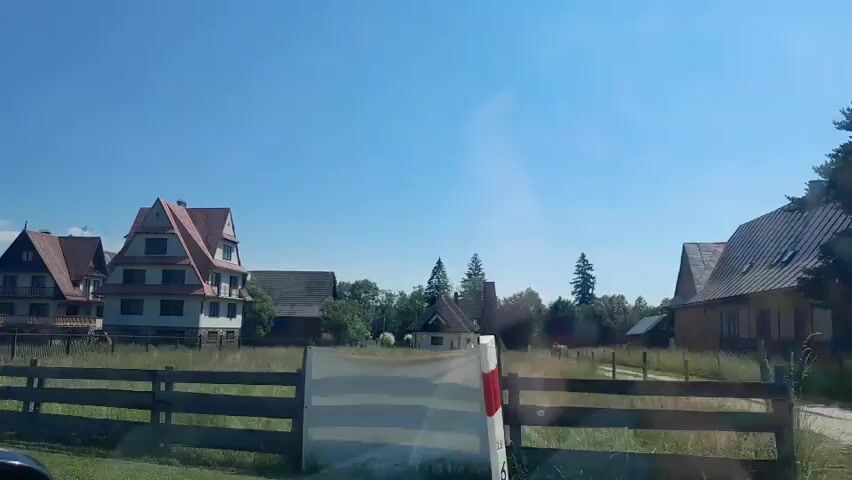 The video shows a rural scene with several houses and trees under a clear blue sky. In the foreground, a wooden fence runs horizontally across the frame. Beyond the fence, a grassy field is visible, with some of the grass appearing dry and yellow. Several houses are scattered throughout the scene, with architectural styles varying from traditional to more modern. One prominent house in the center has a distinctive triangular roof and multiple stories. Another house to the left also features a steep, angled roof. In the background, a line of trees stands out against the sky. The overall impression is a sunny day in a quiet, possibly European, countryside setting. There are no people visible in the video, and the scene appears still and peaceful. The camera appears to be moving slowly, possibly from inside a vehicle, indicated by a slight blur and reflection on the right side of the frame.
