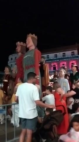 The video shows an outdoor nighttime event. In the foreground, several people are standing in a crowd, some looking at the camera. To the left and center, there are two very large figures, resembling giants, dressed in historical costumes and wearing crowns. These figures appear to be part of a parade or festival. Behind the giants, more people are visible, as well as a building decorated with numerous bright lights. The atmosphere suggests a celebration or cultural event.