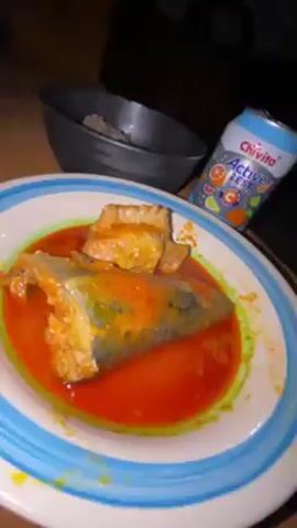 The video shows a plate of fish in red sauce, a bowl of what appears to be pounded yam, and an opened can of Chivita Active Zest drink on a wooden table. The lighting is dim, and there is some movement in the video, suggesting it was taken with a handheld device. A baby voice is heard in the background.