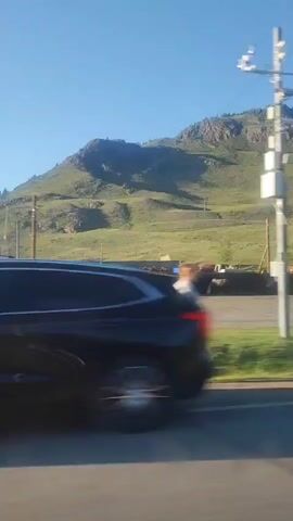 The video captures a view from a moving vehicle. In the foreground, the side of a white car is visible, slightly blurred due to motion. The mid-ground shows a road or paved area, with green grass and a slightly blurry metal fence. Beyond the fence, there appears to be an industrial site with various structures, including a tall, rusty chimney stack and some silo-like buildings. These structures are somewhat weathered and show signs of disuse. In the background, a large, green, grassy mountain with rocky outcrops dominates the landscape under a clear blue sky. The sun appears to be low, casting long shadows and illuminating the scene.