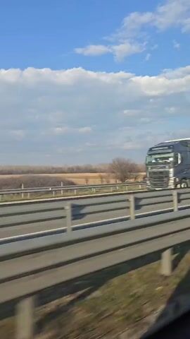 The video shows a view from a moving vehicle on a highway. The sky is blue with scattered white clouds. On the other side of the highway's metal barrier, there is a flat, grassy field with some trees in the distance. There are no people visible in the video. The focus is on the landscape as seen from the vehicle. The overall tone is neutral and observational.