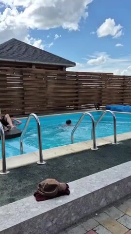 Three people are in a swimming pool. One person is on a lounge float in the shallow end, two other people are in the deeper end of the pool swimming. The pool is surrounded by a wooden fence and has metal steps. There is a small gazebo-like structure behind the fence. The sky is blue with white clouds.