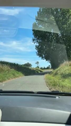 The video shows a view from inside a car traveling on a paved road through a rural landscape. The sky is blue with scattered clouds. On the left side of the road, there are fields with tall green grass. On the right side of the road, there are trees and more grassy slopes. The road curves to the left as it heads towards a group of trees in the distance. The interior of the car is visible in the foreground, including the dashboard and a portion of the windshield with reflections. There are no people or other vehicles visible in the shot. The overall impression is of a scenic drive through the countryside on a clear day.