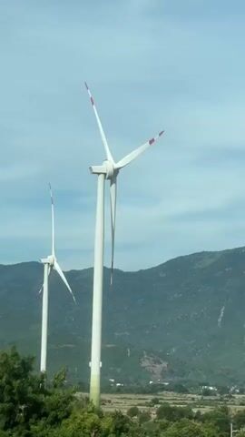 The video displays a landscape featuring two prominent wind turbines situated in Thuan Bac, Vietnam. The turbines, tall and white with red accents on their blades, are positioned against a backdrop of rolling green hills. The sky above is a light blue with scattered clouds, suggesting a partly cloudy day. In the foreground, lush green trees and foliage are visible, adding depth to the scene. The wind turbines are actively generating power, with their blades slowly rotating, indicating a breezy condition. The overall atmosphere is serene and captures a moment of natural energy production within a scenic environment. There is no visible text in the media.
