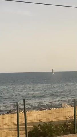 A sailboat is visible on the ocean. In the foreground, there are bushes and a metal fence. The camera pans to the right, revealing more of the ocean and shoreline. Two people are fishing near the water. A sign with a speed limit of 10 is visible.