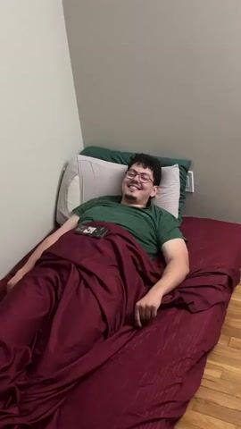 In a casual indoor setting, a man with dark curly hair and glasses is seen lying in bed. He is wearing a green t-shirt and is covered by a burgundy comforter that reaches up to his waist. Behind him, a white pillow rests against a dark green pillow, which is propped against a light grey wall. The floor is made of wood.

The man's smartphone is placed on the comforter next to him. Initially, he forms a "camera" gesture with his hands, holding his index fingers and thumbs up to his eyes as if framing a shot. He then drops his hands, smiles broadly, sticks out his tongue, and laughs playfully, conveying a sense of amusement and lightheartedness. The overall atmosphere is relaxed and informal.
