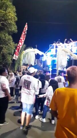 At night, a group of people is gathered for an event. Many of them are wearing white shirts with a design on the back. Some women wear patterned sarongs. A large, decorated structure sits on a platform with speakers and bright lights. People are on the platform. A flag is visible on the left side of the scene. Trees are nearby. The crowd appears to be watching and listening to something happening on the platform.
