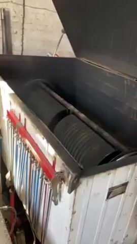 The video shows a large, open, box-like machine in a warehouse-like setting. The machine's interior is dark and contains two large, black, ridged, cylindrical objects rotating on a frame. The machine is white with a black interior and a red accent on the front edge. A row of multi-colored tubes hangs along the front of the machine. The lid of the machine is propped open, revealing the inside. The background shows a white brick wall and a concrete floor. There are no people visible in the video. The primary action is the rotation of the cylindrical objects within the machine. The overall environment appears industrial.
