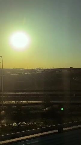 A vertical video captures a view from a moving vehicle on a sunny day. The scene shows a highway with multiple lanes and overpasses, with cars driving in both directions. The sun is bright and low in the sky, casting long shadows and creating lens flare. The landscape is arid, with dry grass and dirt slopes. A traffic light with red lights is visible on a pole. The video pans slightly to the right, revealing more of the highway infrastructure and the surrounding environment. The overall atmosphere is one of travel and urban infrastructure under a bright sky.
