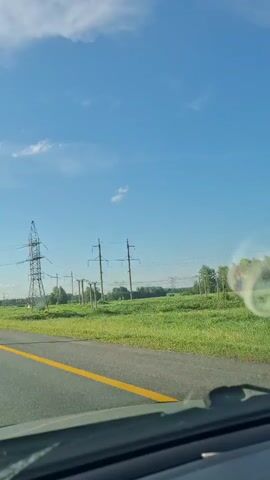 A POV video shows a car driving on a paved road with a yellow line down the center. To the right of the road is a field of green grass and several tall power lines with structures supporting them. The sky is blue with some white clouds. The video appears to be shot during the daytime.