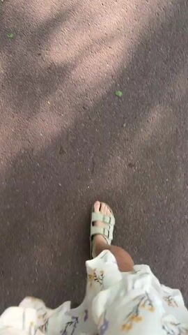 The video begins with a close-up shot of a person walking on a paved surface. They are wearing a white dress with a floral pattern and light-colored Birkenstock sandals. A turquoise ankle bracelet is visible on their left ankle. The camera angle is low, showing mostly the person's legs and feet. The footage then transitions to a wider shot of a road running through a dense forest. The trees are lush and green, creating a canopy of leaves overhead. A "no entry" sign is visible on the right side of the road. The camera pans slightly to show more of the surrounding forest.