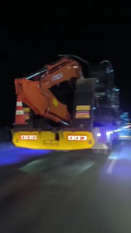 A low-angle nighttime shot captures the side of a flatbed truck as it travels on a road. The truck is carrying an orange Hitachi excavator. The excavator's arm and bucket are clearly visible. The truck has yellow and purple accent lights. The road has yellow and white lane markings, and other vehicles and distant lights are visible in the background. The camera appears to be moving alongside the truck.