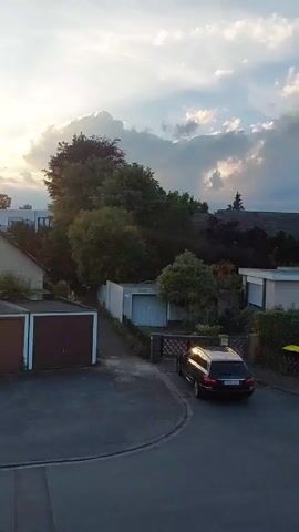 Here is a description of the video:

The video showcases a residential scene during sunset or sunrise. The camera seems to be positioned at an elevated viewpoint, like a balcony or a high window, overlooking a row of garages and houses. 

The foreground features several garages. Some have plain brown doors, while one has a colorful mural depicting cartoon bees. Further along, another garage door is adorned with artwork showing an astronaut character standing on a pink cartoon animal on a planet. 

Behind the garages is a mix of single-family homes, each with varying designs and colors. Some have well-maintained gardens, and trees are interspersed throughout the scene. The sky is bright, with the sun partially obscured by clouds, casting a warm glow over the landscape.