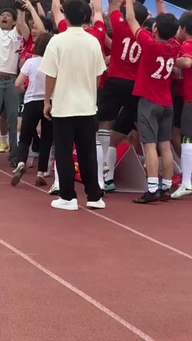 A group of people, including soccer players in red jerseys, are celebrating a victory on a red track. They are holding a trophy and raising their arms in the air. Some people are cheering and clapping. A few people are standing around the group, observing. In the background, there is a building with a sign in Chinese characters. The weather appears to be overcast. The scene is celebratory and joyful.