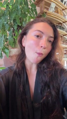 A woman sits in front of the camera in an indoor setting and prepares to eat a salad from a white bowl. She wears a black robe and has long, dark wavy hair. The salad contains various ingredients including cauliflower, tomatoes, yellow peppers, and greens. Behind her, a large leafy plant is visible, along with a decorative structure resembling a multi-tiered display. The woman looks at the camera and then at her salad, picking up a fork to eat.