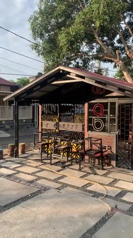 The video shows a small, open-air coffee shop with a covered seating area. The shop has a rustic design with a metal roof and brick accents. There are several tables and chairs set up outside for customers. The ground is paved with rectangular stones and gravel. A large tree with green leaves provides shade over the area. The overall impression is one of a casual and relaxed cafe.