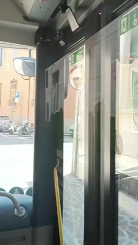 The video shows a view from inside a vehicle, looking out through glass doors. The doors are reflecting the interior of the vehicle, as well as the exterior street scene. The exterior scene consists of buildings lining a street with parked cars. There is a stop sign painted on the road. The sky is blue and clear. The video captures reflections on the glass doors of various objects, including emergency exit signs.