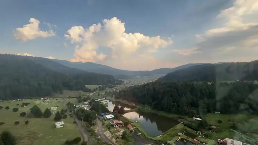 The video shows a scenic view of a valley from a high vantage point, possibly a cable car or gondola. The valley is nestled between tree-covered hills or mountains under a blue sky with scattered clouds. A small lake or pond is visible in the center of the valley, reflecting the sky. A few buildings are scattered around the lake and along a road that winds through the valley. A flash of light reflects off the window. Someone says, "Señor, aqui está padre. Ay, tiene muchas nubes."