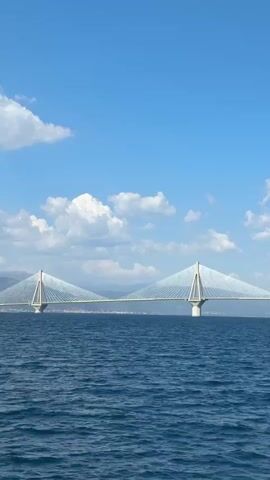 The video captures a breathtaking view of the Rio-Antirrio Bridge, a modern cable-stayed bridge spanning the Gulf of Corinth, connecting the Peloponnese peninsula with mainland Greece. The bridge is the primary subject, its impressive white pylons and delicate network of cables stretching across the wide expanse of blue water. The scene is set under a bright, clear blue sky dotted with fluffy white clouds, suggesting a pleasant, sunny day.

The perspective is from a boat moving across the water, evidenced by the slight rocking motion of the frame and the glimpse of a metal railing in the foreground. The water is a deep, vibrant blue, with visible ripples indicating a gentle breeze. In the distance, the coastline of Greece is faintly visible, with rolling hills and a hint of the urban landscape of Rio.

There are no people visible in the media. The activity is the passive observation of the majestic bridge from the water. The time of day appears to be mid-morning or early afternoon, given the bright sunlight and the shadows cast by the bridge. The overall mood is one of awe and admiration for the engineering marvel and the natural beauty of the setting. The location cues, the iconic bridge itself and the visible coastline, firmly place the scene in the Municipal Unit of Rio, Greece. No text is visible in the media.