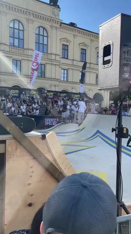A group of young skateboarders, some wearing helmets, stand on a ramp structure with a crowd watching from bleachers in the background. A man in a white t-shirt and beige pants, likely an announcer or organizer, is handing out awards or medals to the skateboarders. The ramp has yellow and blue striped designs. Flags with "SM" and "SVENSKA SPEL" are visible. The scene appears to be an outdoor event in a city with a building in the background.