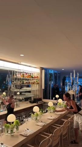 The video shows the interior of a bar or restaurant. In the foreground, a woman in a black tank top and white striped shorts is standing at a bar, holding a bunch of green flowers. She is looking towards her left. The bar is set with place settings, small lamps, and more green floral arrangements. Behind the bar, shelves are stocked with bottles of alcohol and glassware. In the background, two men in suits appear to be talking near a window. The overall lighting is warm and ambient. Another woman, partially visible on the left, is walking away from the camera.