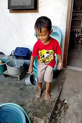 A young boy wearing a red t-shirt with a cartoon character and patterned shorts is playing with water. He is standing on a wet concrete surface outside. In his right hand, he holds a metal ladle, from which he is pouring water onto the ground. His feet are bare and wet. To his left, there are several objects, including a large metal pot on a stand and a blue plastic container. Behind him, there is a large, broken blue plastic bathtub. The background shows a white wall, a doorway, and shelves with various items. The boy is looking down at the water he is pouring.