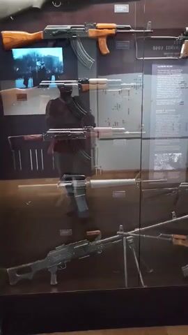 The video displays a museum exhibit featuring a variety of firearms. The firearms are arranged vertically in several rows behind a glass barrier. The exhibit appears to be well-lit, highlighting the details of each weapon. In the reflection on the glass, a person is visible wearing dark clothing. The background of the exhibit includes informational plaques with text and images, suggesting a historical or educational context. Some of the visible firearms include what appear to be AK-47 variants, an M16 variant, and a machine gun. The overall scene is static, with no apparent movement other than the slight camera shake.