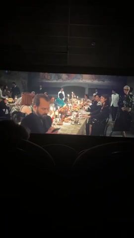 The video shows a movie screen in a darkened cinema. The scene on the screen depicts a formal dinner taking place beneath a mural of The Last Supper. A long table is laden with food, candelabras, and glassware.  People are seated around the table, dressed in formal attire. An older woman with short white hair and glasses, wearing a sequined jacket, gestures towards the table. A man in the foreground is looking at his phone. The rows of empty cinema seats are visible in the foreground.