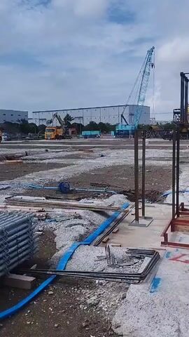 A construction site is depicted. In the foreground, rebar and concrete forms are visible. A metal cage-like structure is being assembled, with a small crane attached. In the background, a large warehouse-style building and several power transmission towers are present. Various construction vehicles, including a yellow excavator and trucks, are scattered across the site. The sky is cloudy.