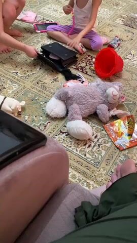 A group of people, mostly children, are gathered on a patterned carpet. In the foreground, a person's legs are visible, with a tablet on a pink chair. In the mid-ground, a girl in a white tank top and purple pants is rummaging through a black bag. Next to her is a stuffed gray cat lying on its side, and an orange snack bag. A red bucket is also on the floor. The overall impression is a casual indoor gathering, possibly a play session, with toys and snacks scattered around.