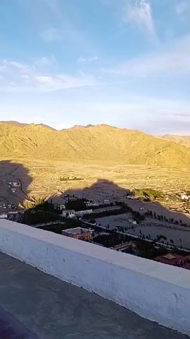 The video displays a sweeping aerial view of a valley with mountains in the background. The landscape is dotted with buildings, trees, and roads, suggesting a populated area. The lighting indicates it is either sunrise or sunset, casting long shadows across the terrain. The sky is partly cloudy. The camera appears to be positioned on a raised platform, with a white railing in the foreground.