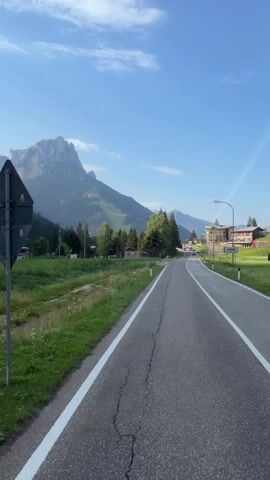 This is a first-person perspective video of a person on a road. The road is paved and has white lines on the side. To the left of the road, there is a grassy area with some trees and a mountain in the background. There are also some vehicles parked on the left side of the road. To the right of the road, there are some buildings and more grassy areas. The sky is blue with some clouds. The video appears to be taken during the daytime.